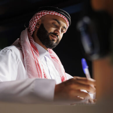 Detailed image showcasing focused Arab entrepreneur at the table writing on a notebook. Close-up shot of a Muslim man using a pen for taking research notes in his home office.