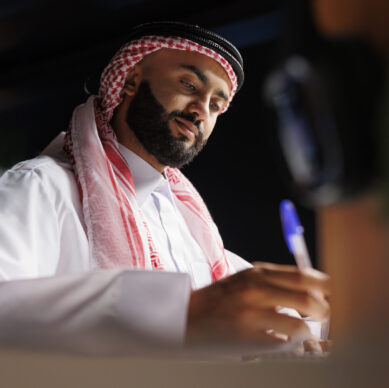 Detailed image showcasing focused Arab entrepreneur at the table writing on a notebook. Close-up shot of a Muslim man using a pen for taking research notes in his home office.