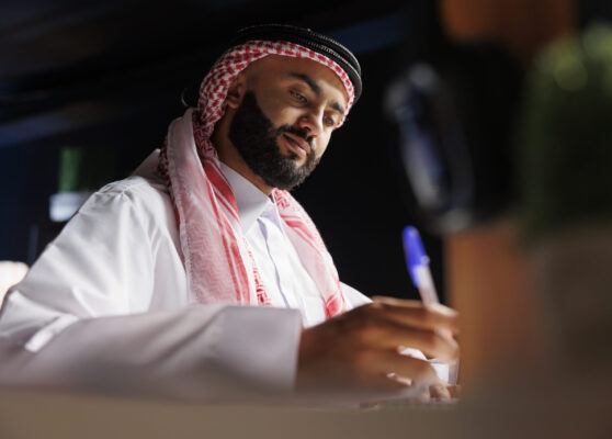 Detailed image showcasing focused Arab entrepreneur at the table writing on a notebook. Close-up shot of a Muslim man using a pen for taking research notes in his home office.